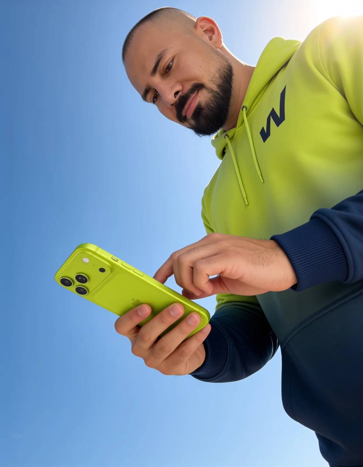 Un hombre con sudadera verde mira la pantalla del teléfono que sostiene con las dos manos, fotografiado desde abajo contra un cielo azul.