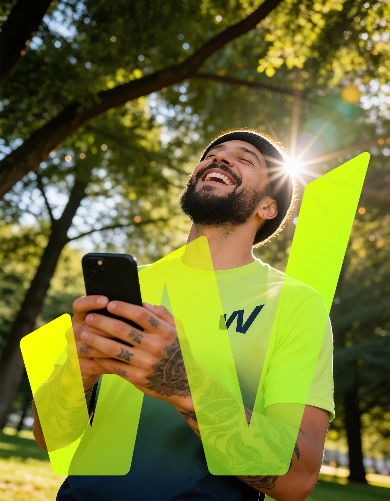 Un hombre sonríe con la cabeza levantada hacia el sol, sosteniendo un teléfono, con la luz filtrándose entre los árboles de un parque.